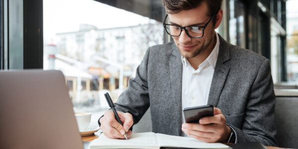 Smiling business man in eyeglasses sitting by the table in cafe with laptop computer while using smartphone and writing something